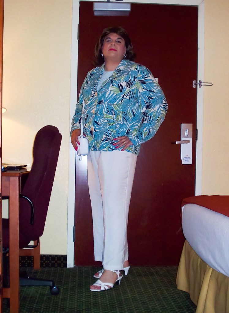 A person posing confidently in a hotel room, wearing a colorful floral jacket, white pants, and sandals, standing near a door.