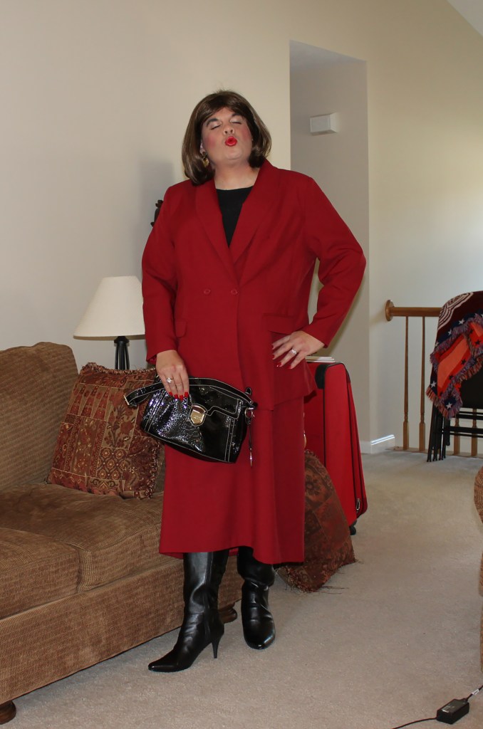 A person wearing a red suit with a black top and knee-high black boots, posing in a living room with a beige sofa and a suitcase nearby.