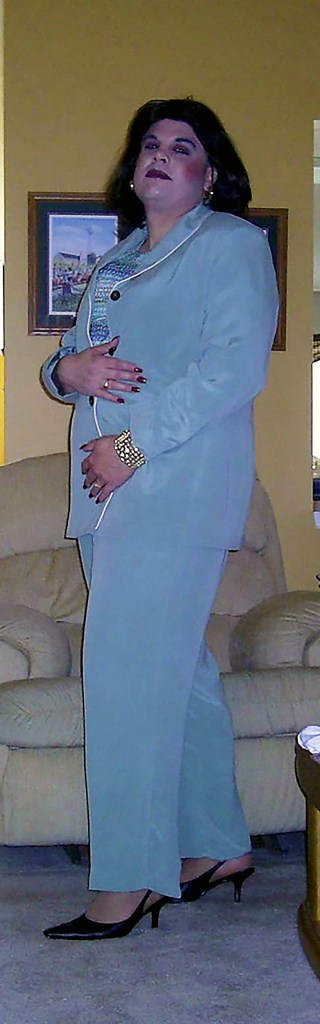 A person wearing a light blue suit with a stylish blouse, standing confidently in a living room setting, with a beige couch in the background.