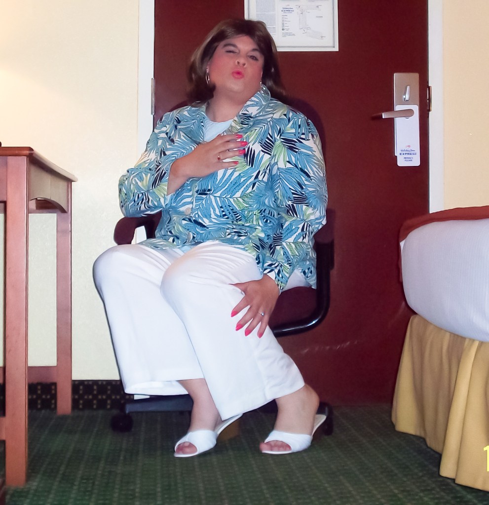 A person sitting in a chair in a hotel room, wearing a colorful tropical print jacket, white pants, and sandals, with a playful expression and hand on their heart.