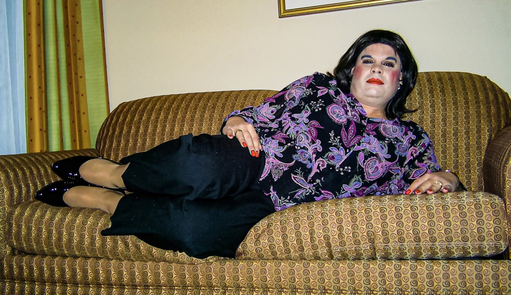 A person lounging on a patterned couch, wearing a colorful blouse and black pants, with makeup and styled hair.
