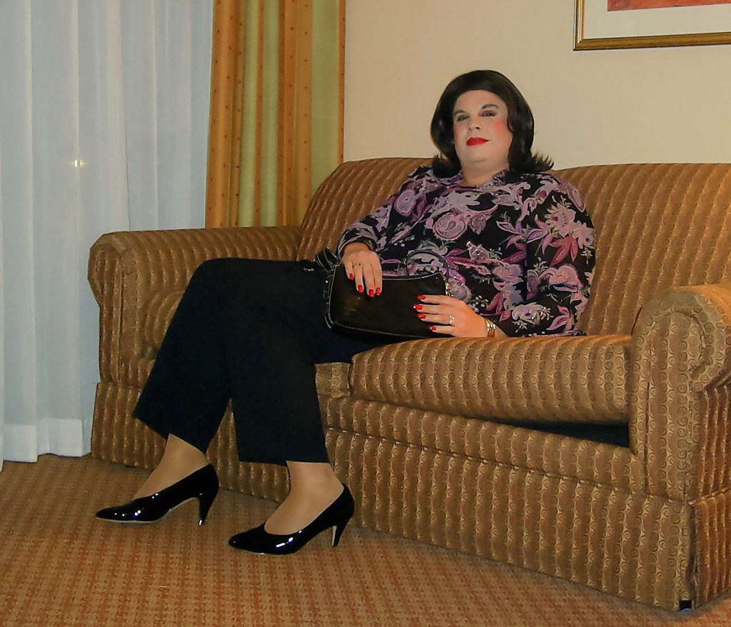 A person sitting on a patterned sofa, wearing a floral top and black pants, accessorized with black heels and holding a small handbag.