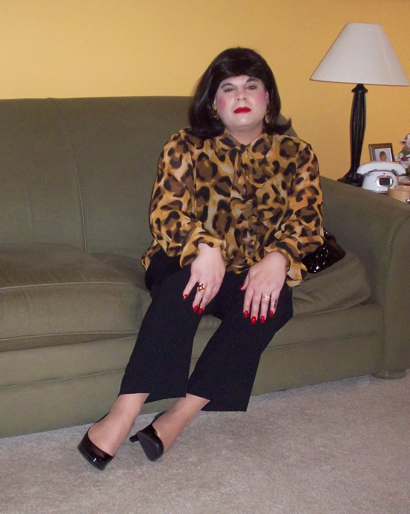 A person sitting on a green couch, wearing a leopard print blouse and black pants, with red nail polish and black high heels, in a room with a yellow wall and a lamp.