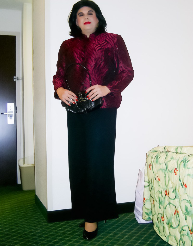 A person dressed in a burgundy textured jacket and long black skirt, holding a black handbag, posing in a well-lit indoor setting with a green carpet.
