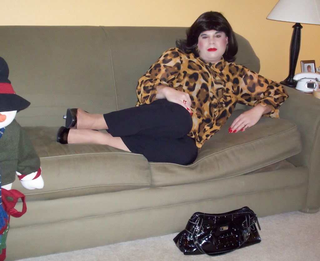 A person lounging on a green couch, wearing a leopard print blouse and black pants. They have dark hair and red lipstick, with manicured nails. A black handbag is placed next to them, and a decorative snowman figure is visible in the foreground.