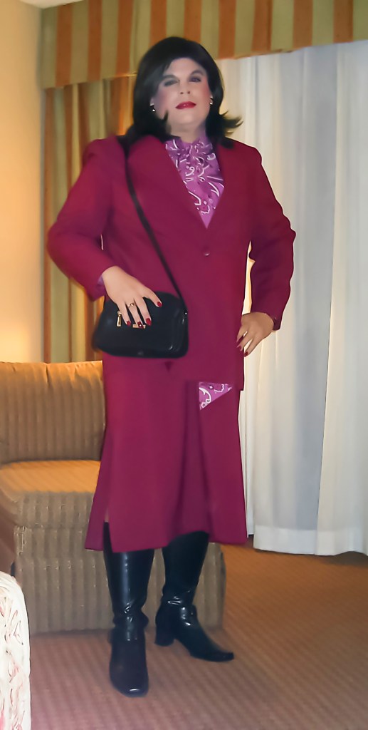 A person stands confidently in a hotel room, wearing a tailored maroon suit with a purple blouse. They hold a small black handbag and are wearing knee-high black boots. The background features striped curtains and a beige couch.
