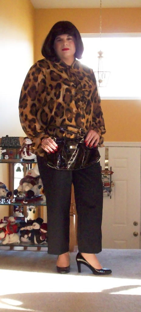 A person standing indoors, wearing a leopard print blouse, black pants, and high heels, with a shiny black purse. The background features a shelf with decorative items.