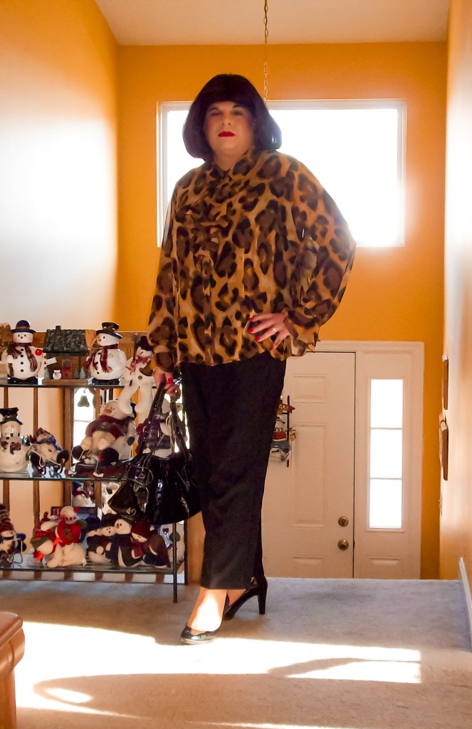 Person in a leopard print blouse and black pants standing in a brightly lit hallway with yellow walls, holding a black handbag.