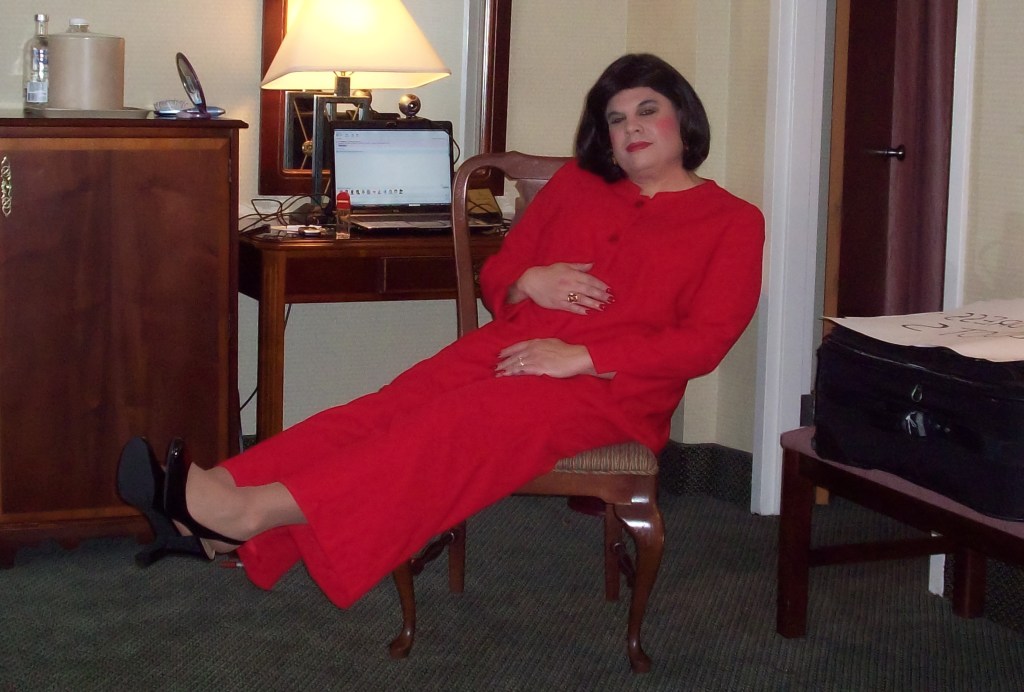 A person sitting casually in a hotel room, wearing a red outfit and black heels, with a relaxed expression. A desk with a laptop and other items is in the background.