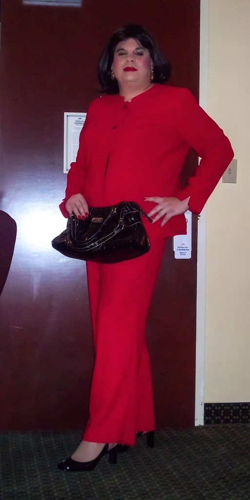 A person in a red suit stands in front of a wooden door, holding a black handbag. The individual has long dark hair and is wearing black high-heeled shoes.