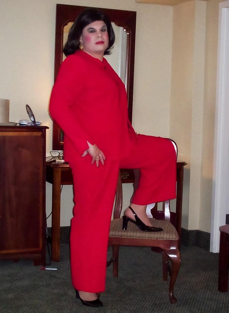 A person in a striking red suit poses confidently, with one leg resting on a chair, wearing black high heels in a well-lit indoor setting.