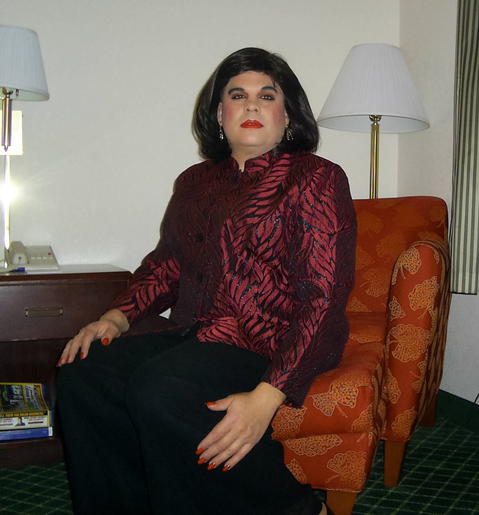 A person sitting on an orange patterned chair, wearing a red and black patterned blouse. They have long dark hair, bold makeup, and red nail polish. A lamp and a nightstand are visible in the background.